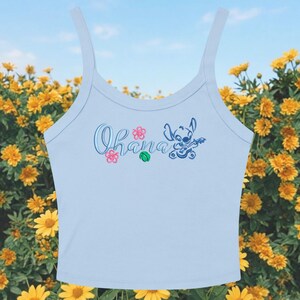 May include: Light blue tank top with the word "Ohana" in script, along with a cartoon character playing a ukulele. Pink and green floral accents are also present. The top has thin straps and a scoop neckline.