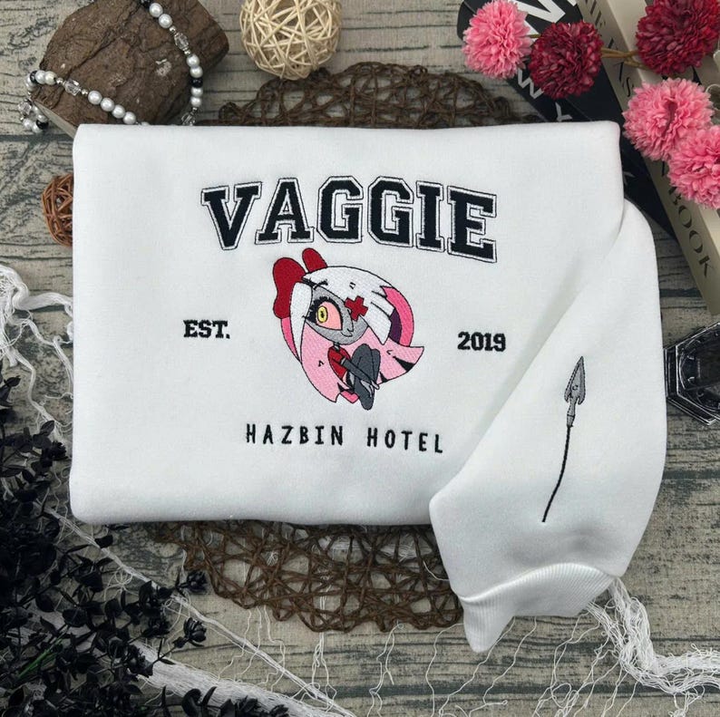 May include: White sweatshirt with the word "VAGGIE" in black block letters, a cartoon character graphic, and the text "HAZBIN HOTEL". The sleeve features a black spear design. The sweatshirt also has the text "EST. 2019".