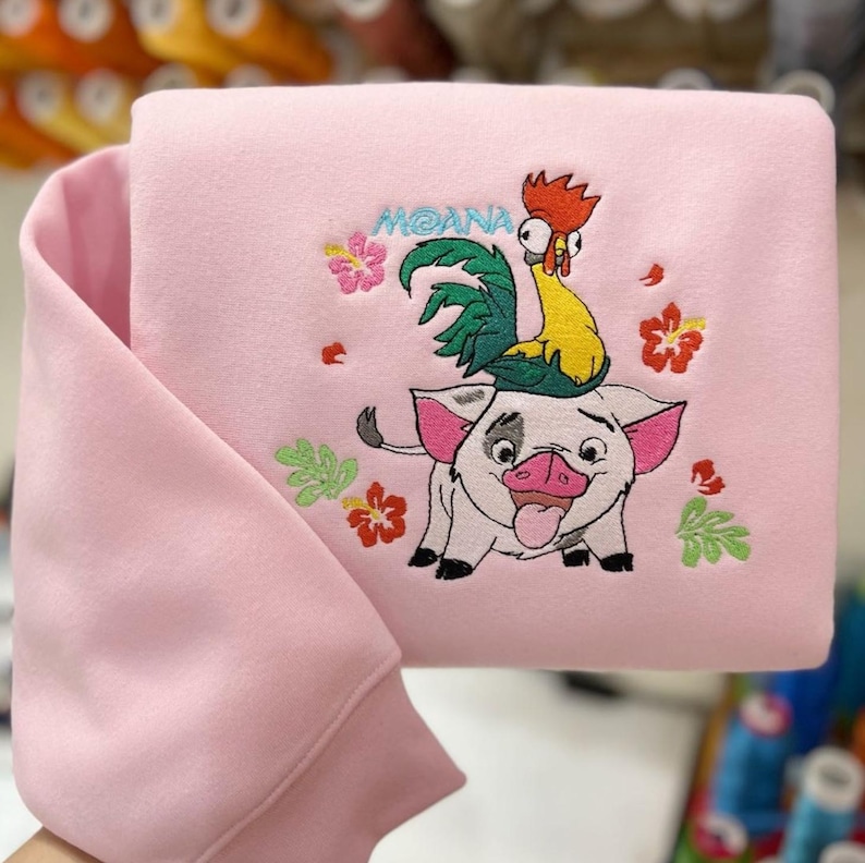 May include: Pink sweatshirt featuring an embroidered design with the word "MOANA" above a cartoon pig with a rooster on its back. The design includes colorful flowers and leaves. The sweatshirt has a folded sleeve.