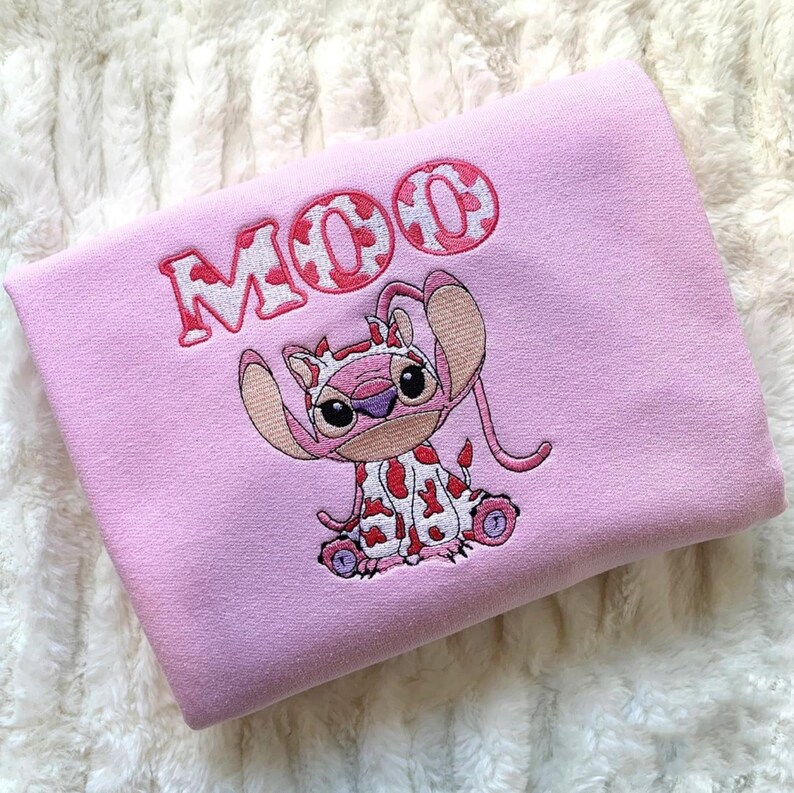 May include: A light pink sweatshirt featuring an embroidered graphic of a character wearing a cow-print hood and outfit. Above the character, the word MOO is embroidered in a matching cow-print pattern with a red outline on the fabric surface.