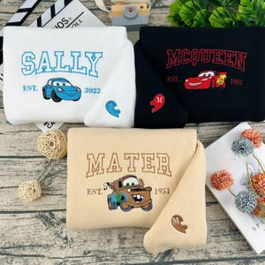 May include: Four embroidered sweatshirts in white, black, and beige. The white sweatshirt says "SALLY" with a blue car graphic. The black sweatshirt says "MCQUEEN" with a red car graphic. The beige sweatshirt says "MATER" with a brown car graphic.