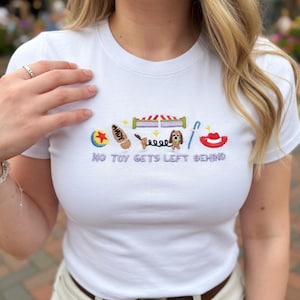 May include: White t-shirt with embroidered Toy Story-themed graphics. The design includes a ball, Woody's name, a bed, Slinky Dog, a horseshoe, and a red hat. The text "NO TOY GETS LEFT BEHIND" is embroidered below the graphics.