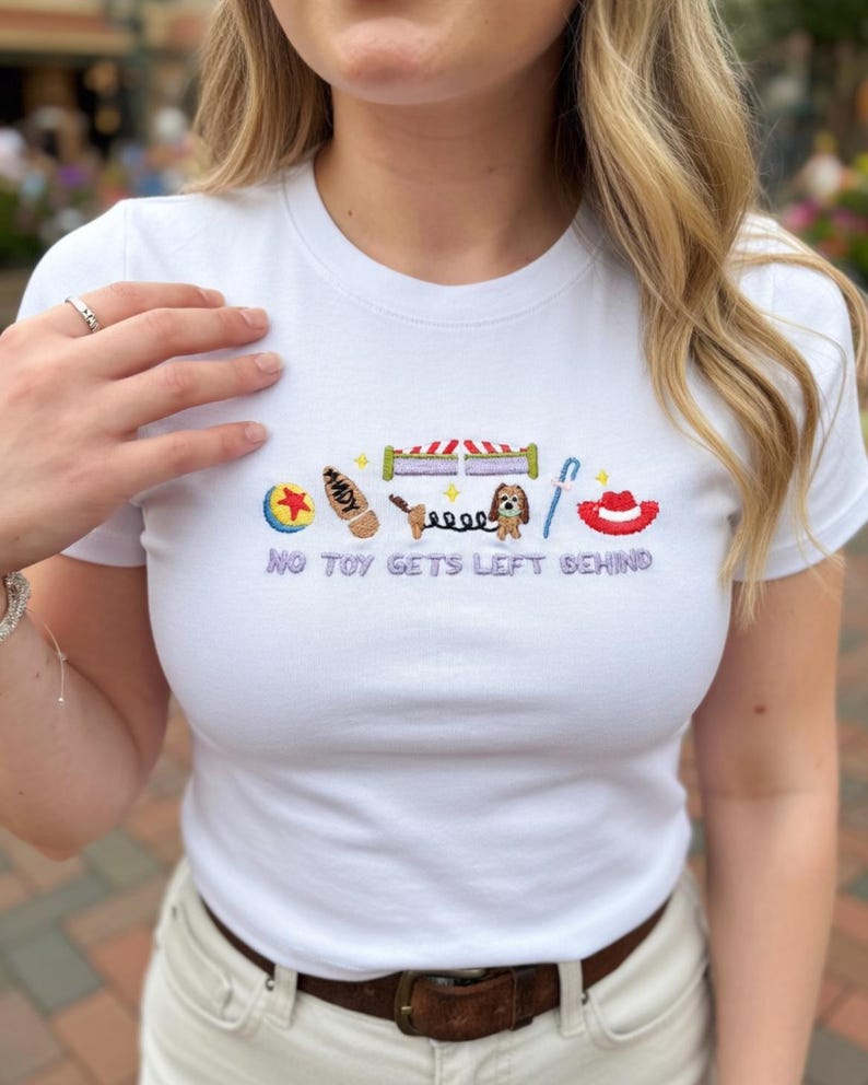 May include: White t-shirt with embroidered Toy Story-themed graphics. The design includes a ball, Woody's name, a bed, Slinky Dog, a horseshoe, and a red hat. The text "NO TOY GETS LEFT BEHIND" is embroidered below the graphics.