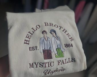 May include: Cream-colored sweatshirt with embroidered text and graphic. The design includes the words "HELLO BROTHER", "EST. 1864", "MYSTIC FALLS Virginia", and two figures. The embroidery is in shades of brown and green.
