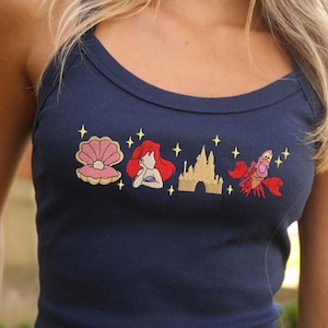 May include: Navy blue vest top featuring embroidered images of a seashell, a red-haired mermaid, a castle, and a red crab. The top has a scoop neckline and is made of a soft material.