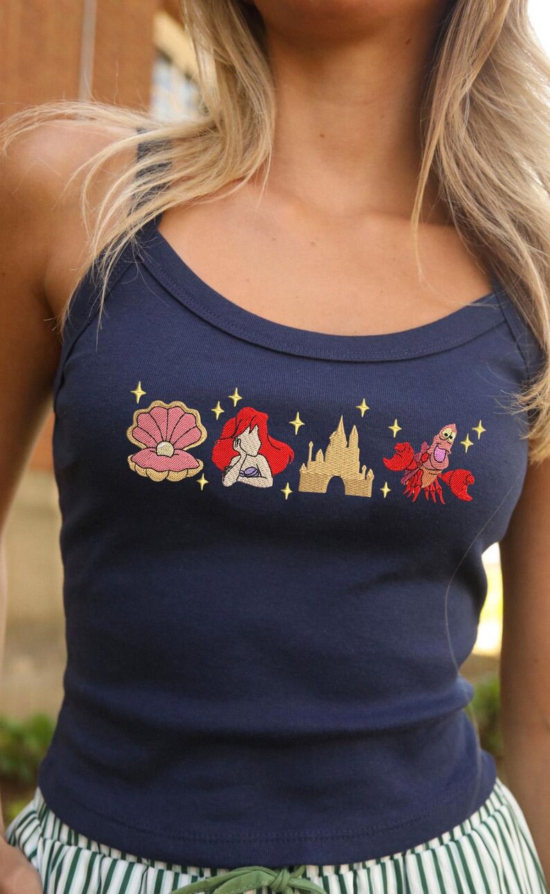 May include: Navy blue vest top featuring embroidered images of a seashell, a red-haired mermaid, a castle, and a red crab. The top has a scoop neckline and is made of a soft material.