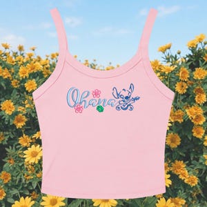 May include: Pink tank top with the word "Ohana" in blue script, along with a cartoon character playing a ukulele. The top has thin straps and is set against a backdrop of yellow flowers and a blue sky.