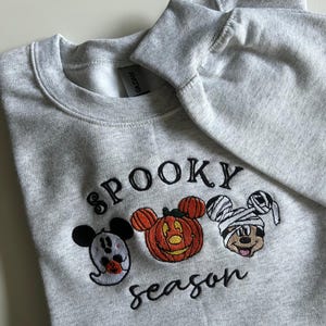 May include: A light gray sweatshirt with the embroidered text "Spooky Season". The design features three cartoon characters dressed as a ghost, a jack-o'-lantern, and a mummy. The sweatshirt is made of a soft material.
