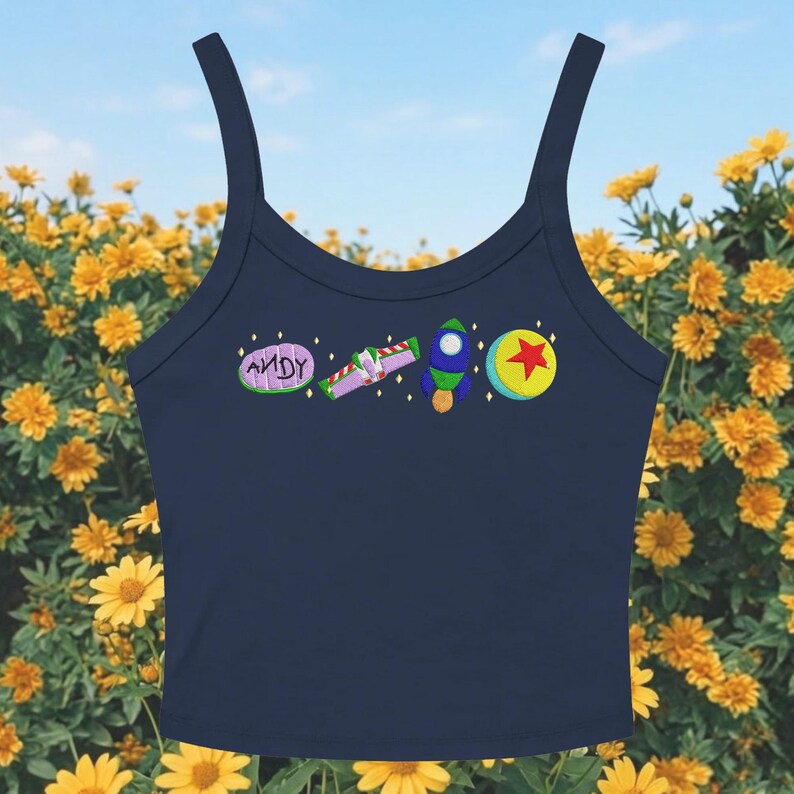 May include: Navy blue crop top with embroidered cartoon character designs. The design includes the word "ANDY", a purple and green ray gun, a rocket ship, and a yellow ball with a red star. The top has a scoop neck and spaghetti straps.