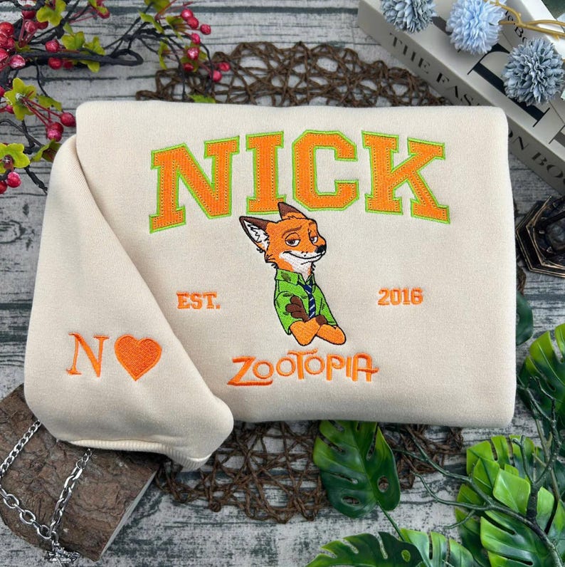Puede incluir: Sudadera color crema con el nombre "NICK" en letras de bloque naranja con ribete verde y un personaje de zorro de dibujos animados. La sudadera tambi&eacute;n tiene el texto "EST. 2016" y "ZOOTOPIA". La manga muestra una "N" y un coraz&oacute;n.