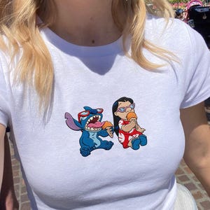 Peut inclure: T-shirt blanc avec un motif color&eacute; de Stitch et Lilo, chacun tenant un cornet de glace. Stitch est bleu avec des lunettes de soleil, et Lilo porte une robe rouge. Le t-shirt a un col rond et des manches courtes.