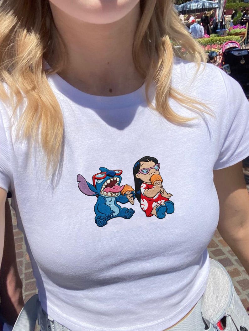 Peut inclure: T-shirt blanc avec un motif color&eacute; de Stitch et Lilo, chacun tenant un cornet de glace. Stitch est bleu avec des lunettes de soleil, et Lilo porte une robe rouge. Le t-shirt a un col rond et des manches courtes.
