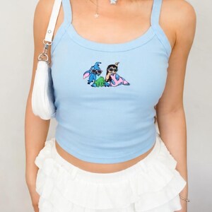 Puede incluir: Camiseta sin mangas azul claro con un dibujo animado de dos personajes. La camiseta tiene tirantes finos y un escote redondo. Combinada con una minifalda blanca con volantes. Un bolso de hombro blanco y un collar de cuentas de colores completan el look.