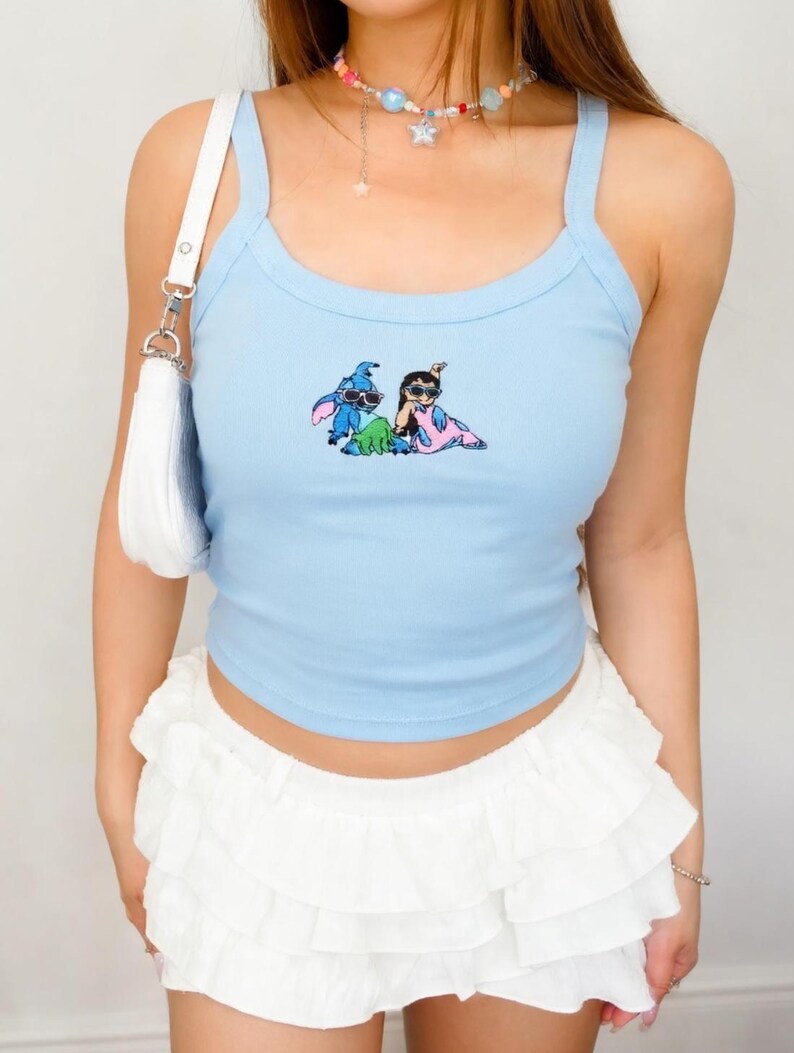 Puede incluir: Camiseta sin mangas azul claro con un dibujo animado de dos personajes. La camiseta tiene tirantes finos y un escote redondo. Combinada con una minifalda blanca con volantes. Un bolso de hombro blanco y un collar de cuentas de colores completan el look.