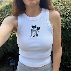 May include: White ribbed tank top with a black and blue embroidered design of a robot character and Mickey Mouse ears. The tank top is sleeveless and has a round neckline.