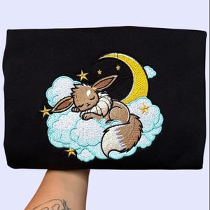 Könnte beinhalten: Schwarzes Sweatshirt mit einem gestickten Design einer schlafenden Cartoon-Kreatur mit braunem Fell, die auf einer Wolke mit einem gelben Halbmond und Sternen ruht. Das Design ist in Braun-, Gelb- und Blautönen gehalten.