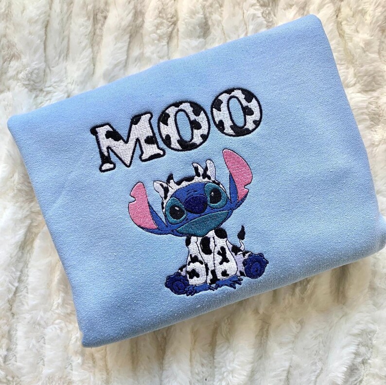 May include: A light blue sweatshirt featuring an embroidered design of a blue alien character with large pink ears and a black and white cow-print pattern on its body. Above the character, the word MOO is embroidered in a black and white cow-print font.