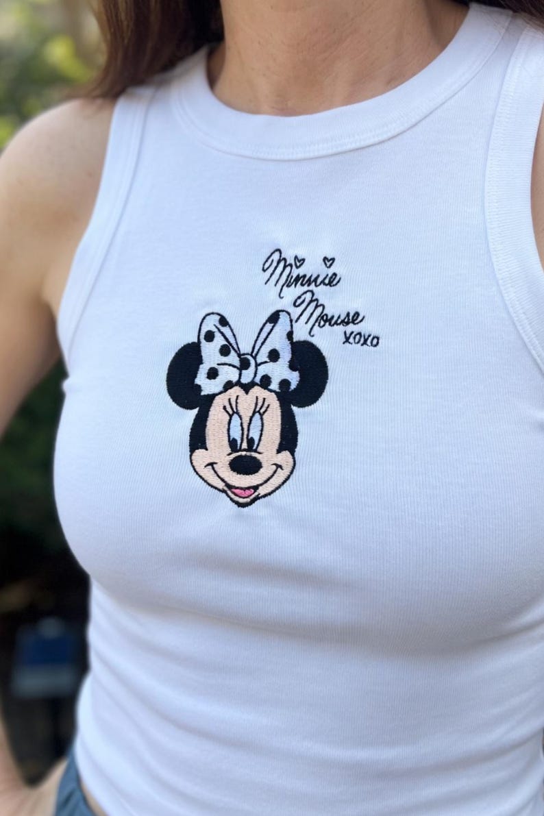 Może przedstawiać: Biały top na ramiączkach z haftowanym wzorem Myszki Minnie. Wz&oacute;r zawiera twarz Myszki Minnie, kokardę w kropki oraz napis "Minnie Mouse XOXO" czarną czcionką. Swobodny top bez rękaw&oacute;w.