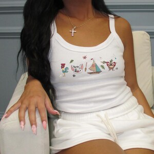 K&ouml;nnte beinhalten: Wei&szlig;es geripptes Tanktop und passendes Shorts-Set. Das Tanktop zeigt farbenfrohe Cartoon-Charakter-Grafiken. Die Shorts haben einen Kordelzug. Eine goldene Halskette mit einem Kreuzanh&auml;nger ist ebenfalls sichtbar.