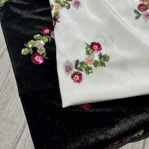May include: Two pieces of velvet fabric, one black and one white, both featuring embroidered floral motifs. The flowers are decorated with small, shiny sequins in shades of magenta, orange, and green, creating a textured, reflective surface.