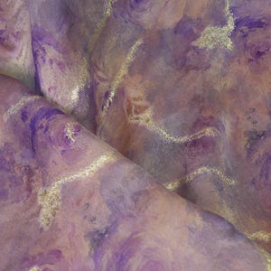 Può includere: Primo piano di un tessuto con effetto acquerello. Il tessuto presenta vortici di tonalità viola, lavanda e pesca, accentuati da linee metalliche dorate. Il materiale appare morbido e fluido, con una leggera lucentezza.