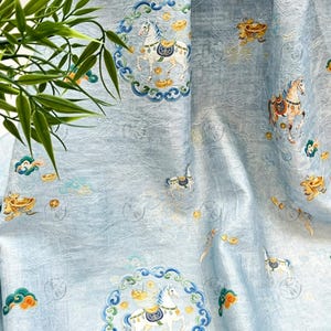 May include: Light blue fabric with an ornate pattern featuring white horses, gold coins, and decorative clouds. The design includes circular frames with detailed borders. The fabric has a textured appearance and a subtle sheen.