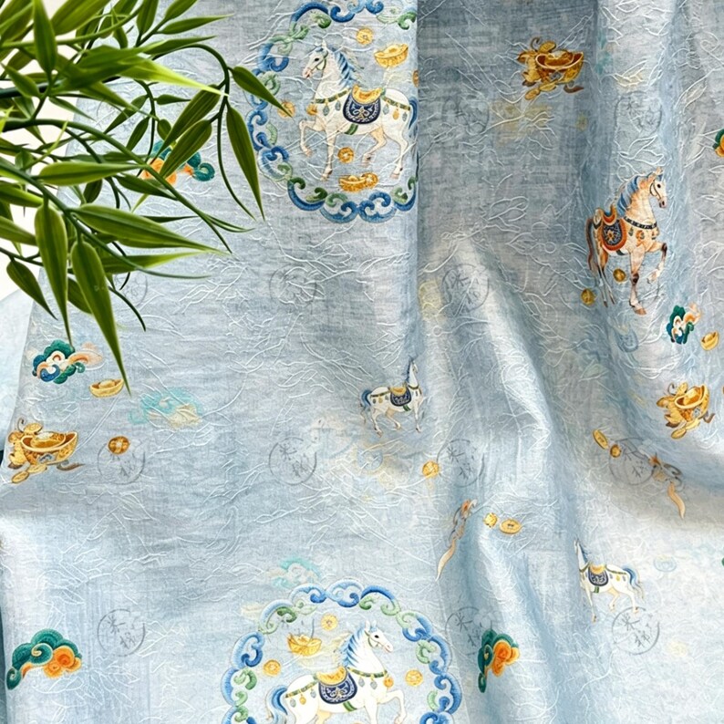 May include: Light blue fabric with an ornate pattern featuring white horses, gold coins, and decorative clouds. The design includes circular frames with detailed borders. The fabric has a textured appearance and a subtle sheen.