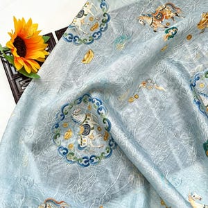 May include: Light blue fabric with an intricate pattern featuring horses, clouds, and decorative circular frames. The fabric has a subtle sheen and a delicate, textured appearance. A sunflower and a decorative wooden frame are in the background.