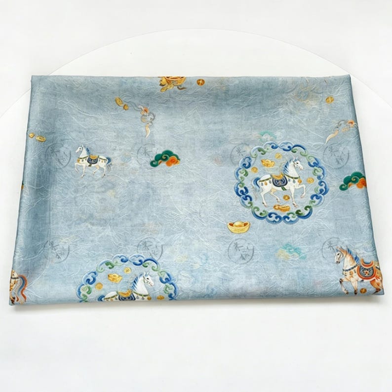 May include: A light blue fabric with a detailed pattern featuring white horses, gold accents, and circular designs. The fabric has a textured appearance and a subtle sheen, with a traditional Asian-inspired aesthetic.