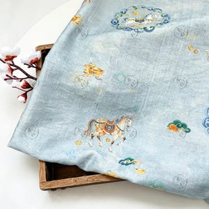 May include: Light blue fabric with a detailed pattern featuring horses, carriages, and clouds in shades of gold, orange, and blue. The fabric has a subtle, textured appearance and is draped over a wooden box.
