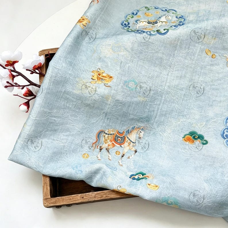 May include: Light blue fabric with a detailed pattern featuring horses, carriages, and clouds in shades of gold, orange, and blue. The fabric has a subtle, textured appearance and is draped over a wooden box.