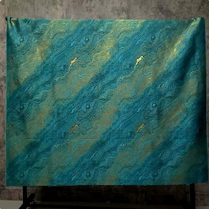 Puede incluir: Tela turquesa y dorada con un patrón veteado, drapeada sobre un soporte negro. El diseño presenta líneas diagonales fluidas y detalles dorados. La combinación crea una apariencia lujosa y artística.