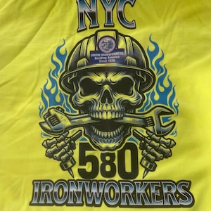 Puede incluir: Camiseta amarilla con un gráfico de calavera negro y azul que lleva un casco. La calavera sostiene dos llaves en su boca. El texto "NYC 580 IRONWORKERS" está impreso debajo de la calavera. Un parche azul y blanco con el texto "UNION IRONWORKERS BUILDING AMERICA Since 1896" está en el casco.