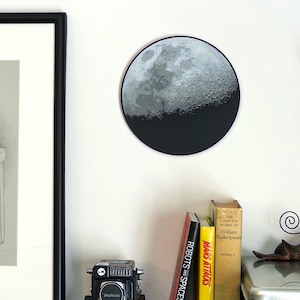 Op de afbeelding: Ronde wandkunst met een gedetailleerd maanoppervlak in grijstinten en een zwart onderste gedeelte. Een vintage camera en boeken staan op een plank eronder. De boeken zijn onder andere 'Robots and Spaceships' en 'Mars Attacks'.