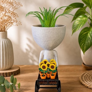 May include: A quirky planter in the form of a figure with a large head, carrying a cart. The head holds a green plant, and the cart contains small pots of sunflowers. The figure and cart are on a wooden surface.