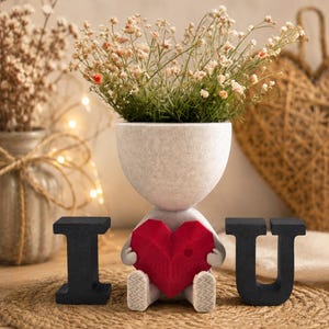 May include: A whimsical planter shaped like a figure holding a red heart, with the letters "I U" beside it. The planter is filled with small flowers. The figure is sitting on a woven mat, creating a charming decorative piece.