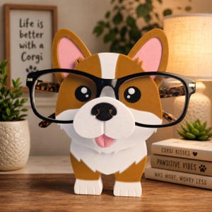 May include: A corgi-shaped eyeglass holder with black-rimmed glasses. The holder is brown, white, and tan with pink inner ears and a smiling face. The text "Life is better with a Corgi" is in the background.