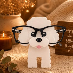Puede incluir: Un soporte para gafas con forma de caniche blanco que presenta un diseño de pelaje rizado y texturizado, ojos negros, nariz negra y una pequeña lengua rosa. La figura lleva puestas unas gafas de montura negra con estampado de leopardo.