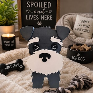 Puede incluir: Una figura decorativa de perro Schnauzer en blanco y negro se encuentra sobre una cama para mascotas de color crema esponjoso, con un juguete de cuerda a juego. Un letrero dice "SPOILED and LIVES HERE". Otros artículos incluyen una vela, una cama para perros, un cuenco que dice "TOP DOG" y un letrero en forma de hueso que dice "RENT FREE".