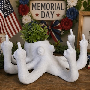 May include: A white octopus-shaped planter with multiple arms, each displaying a raised middle finger. The planter holds green plants and is surrounded by patriotic decorations, including an American flag and a sign that says "ZERO F*CKS GIVEN."