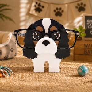 May include: A dog-shaped eyeglass holder wearing black-rimmed glasses. The holder is black, white, and brown, with a cute face. A sign behind the holder says "good dog" and "Wag More Bark Less". A dog bed, toy, and ball are in the background.