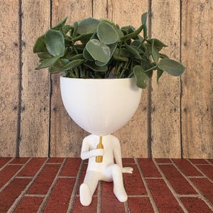 May include: White ceramic planter shaped like a person sitting with a bottle in their hand. The planter has a round head and a large bowl on top for plants. The planter is sitting on a brick surface.