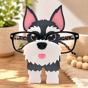 Könnte beinhalten: Ein skurriler Brillenhalter in Form eines Schnauzer-Hundes. Der Halter ist schwarz, weiß und rosa, mit einem verspielten Design. Er zeigt eine Brille mit schwarzen Rahmen und einem Leopardenmuster. Der Halter dient zur sicheren Aufbewahrung von Brillen.
