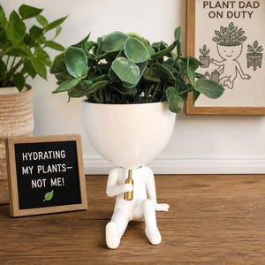 May include: A whimsical white ceramic planter shaped like a person, holding a small gold bottle. The planter is filled with green leafy plants. A framed sign reads "Hydrating my plants - not me!" and another says "Plant Dad on Duty".