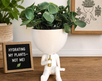 May include: A quirky white ceramic planter in the form of a person, clutching a small gold bottle. The planter is filled with green foliage. A framed sign reads "Hydrating my plants - not me!" and another says "Plant Dad on Duty".