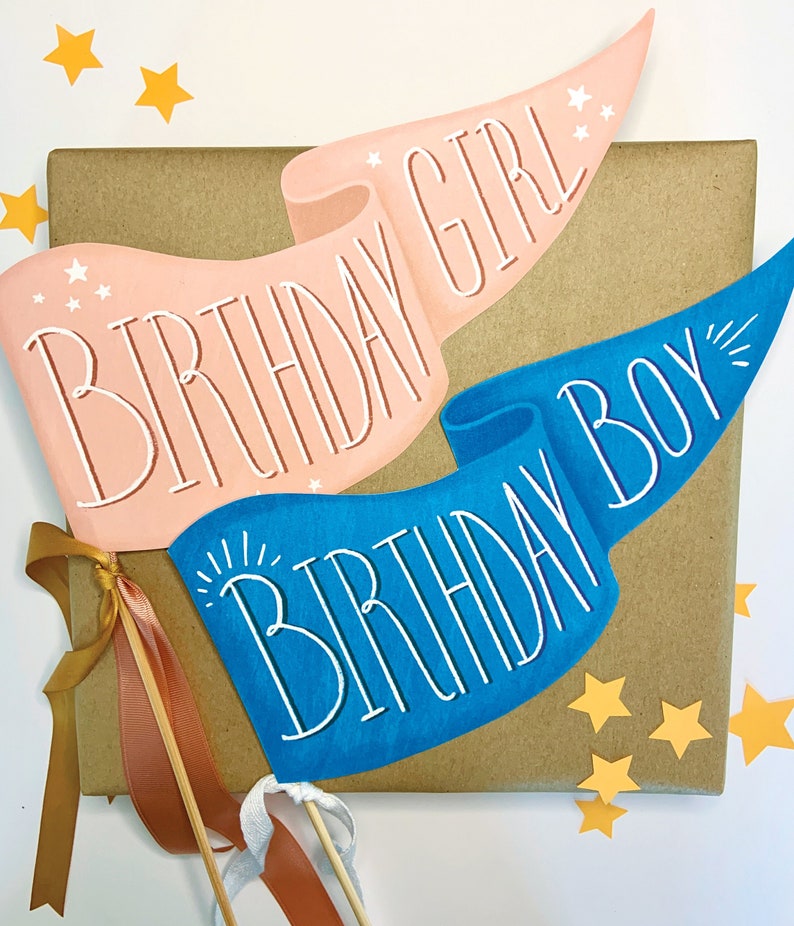 May include: Two paper banners with a birthday theme. The banner on the left is pink with white text that says "Birthday Girl". The banner on the right is blue with white text that says "Birthday Boy".