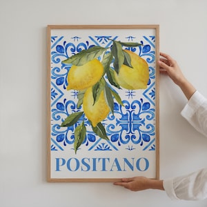 May include: A framed art print with a watercolour illustration of lemons and leaves set against a blue and white patterned backdrop. The word "POSITANO" is printed in blue at the bottom. The frame is made of light-coloured wood.