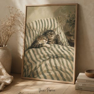 Könnte beinhalten: Ein gerahmter Kunstdruck mit einem Gemälde von zwei getigerten Katzen, die in einem Bett mit grün-weiß gestreifter Bettwäsche liegen. Eine Katze ruht ihren Kopf auf der anderen, während die zweite eine dünne Zigarette hält. Der Rahmen ist aus hellem Holz.