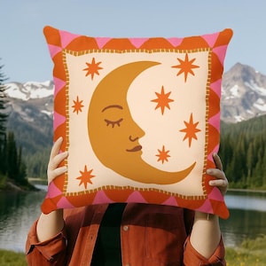 May include: Decorative pillow with a cream-colored background featuring a crescent moon and stars in shades of orange. The pillow has a pink and orange scalloped border. The text "DecorMate" is visible in the background.