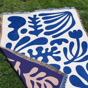 Puede incluir: Una manta tejida reversible con un diseño floral azul y blanco. La manta tiene flecos en todos los lados y un diseño reversible, con un fondo azul y un patrón floral beige en el reverso. Se muestra sobre hierba verde.